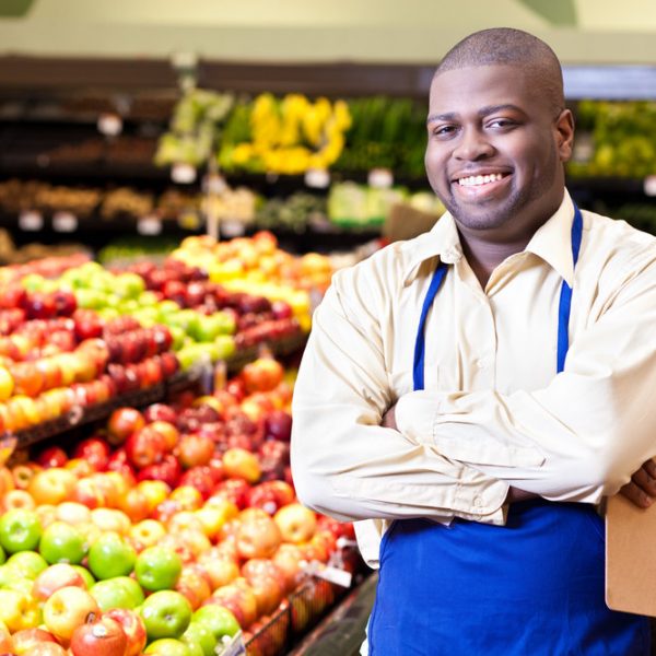 A smiling mid adult man stands in the produce section of his grocery store and smiles for the camera with folded arms.  He wears an apron and carries a clipboard.